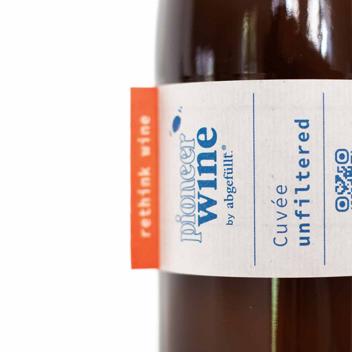 Eine Nahaufnahme der Naturweinflasche 'Unfiltered' von Pioneerwine in einer Mehrwegflasche mit einem wasserlöslichen Etikett. Das außergewöhnliche Design der Flasche und das umweltfreundliche Etikett spiegeln den nachhaltigen Ansatz der Marke wider und machen die Flasche besonders leicht wiederverwendbar.